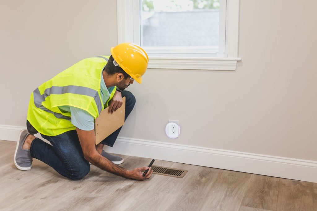 Contractor kneeling to inspect a floor supply vent inside a home